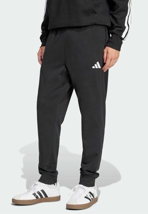 Persona con pantalones de chándal negros de Adidas y zapatillas blancas de Adidas con rayas negras, de pie contra un fondo claro y sencillo.