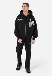 Crna zip-up dukserica s prednjim džepovima, ukrašena šarenim aplikacijama i natpisima. Uparena s crnim hlačama od teksturirane tkanine i crnim tenisicama.