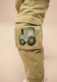 Khaki cargo nadrág, jobboldalon egy texturált zöld és barna hímezett traktorfolttal a térdnél, látható varrási részletekkel.