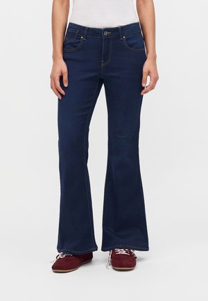 Person iført mørkeblå flared jeans, hvid top og røde sneakers med hvide snørebånd, stående mod en ensfarvet lys baggrund.