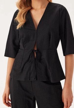 Femme portant un haut noir à manches trois-quarts en V avec fermeture nouée à la taille et pantalon noir assorti.