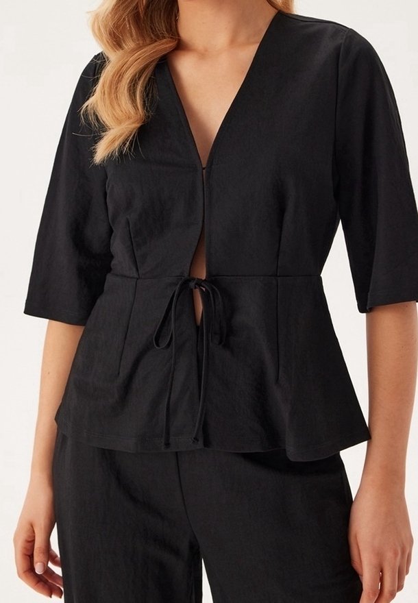 Femme portant un haut noir à manches trois-quarts en V avec fermeture nouée à la taille et pantalon noir assorti.