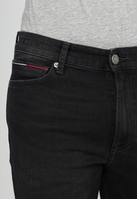 Vaqueros de mezclilla negros con un ajuste slim, que presentan un diseño clásico de cinco bolsillos y un sutil detalle de rayas rojas y azules en el bolsillo delantero.