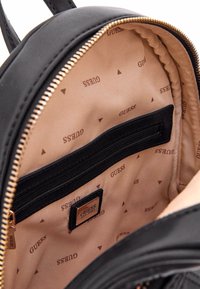 Interno dello zaino nero con fodera beige, presenta il logo del marchio, una tasca con zip e un tessuto a motivi con design del logo ripetuto.