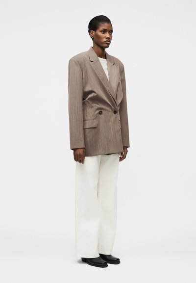 Γυναίκα που στέκεται φορώντας ένα υπερμεγέθες καφέ σακάκι με διπλό πέτο, λευκό παντελόνι φαρδύ στο κάτω μέρος και μαύρα παπούτσια, με φόντο ένα απλό σκηνικό.