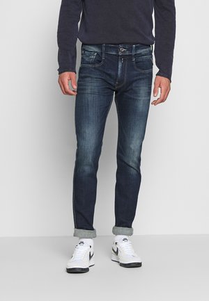 Homme portant un jean slim bleu foncé avec les revers retroussés, des baskets blanches Nike et une chemise sombre à manches longues, debout à l’intérieur.