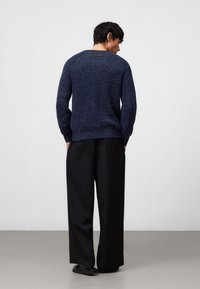 Navyblauer Strickpullover mit strukturiertem Finish, rundem Ausschnitt und gerippten Bündchen. Schwarze weit geschnittene Hosen und dunkle Schuhe runden das Outfit ab.