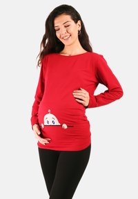 Rotes Schwangerschaftsoberteil mit einem Cartoon-Babymotiv am Bauch, langen Ärmeln und einem lockeren Schnitt. Kombiniert mit schwarzen Leggings.