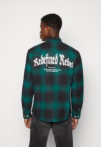 Zielona i czarna flanelowa koszula w kratę z białym napisem na plecach. Długie rękawy, mankiety na guziki i zaokrąglony dół. Miękka tekstura, casualowy design.