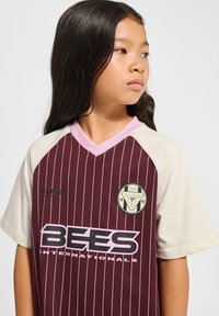 Maron- og beige trøje med lodrette striber, korte ærmer, lyserød krave, "BEES" logo og et rundt mærke med et grafisk design.