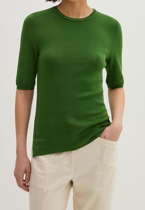 Femme portant un top en maille vert ajusté à manches courtes avec des côtes verticales et un pantalon écru, une main dans la poche.