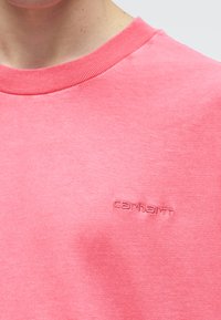 Κοντινό πλάνο ενός ατόμου που φοράει ροζ T-shirt Carhartt με ριμπ ύφανση στον στρογγυλό λαιμό και κεντημένο λογότυπο στο στήθος.