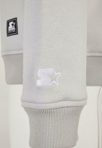 Manche d'un sweat-shirt gris clair avec un "S" brodé en blanc et un logo étoile au-dessus du poignet côtelé, et une étiquette de la marque noire sur la manche supérieure.