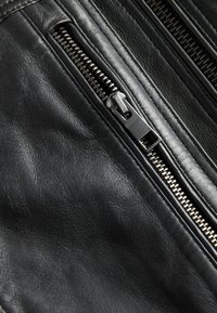 Prenda de cuero negro con cremalleras visibles. La textura parece suave y flexible, mostrando el hardware metálico de las cremalleras en la parte delantera.
