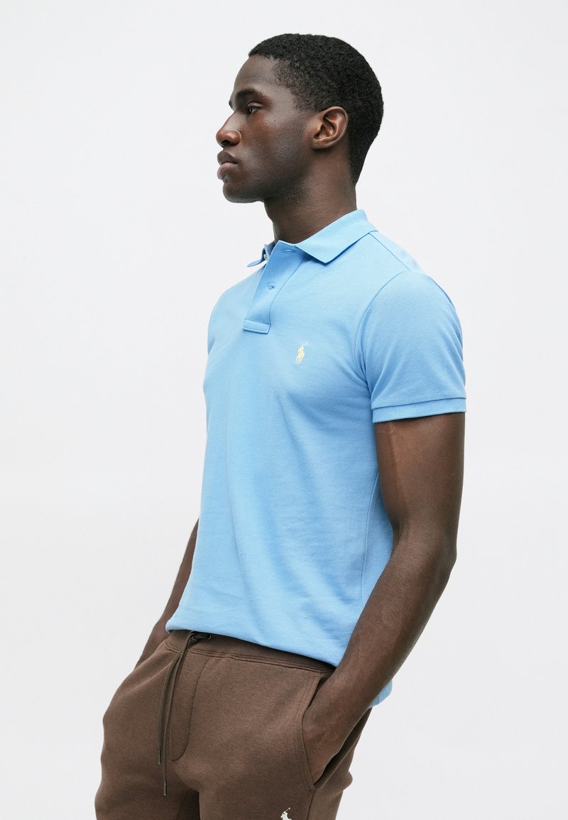 Lichtblauwe polo met een textuurstof, kraag met knopen en een klein geel logo geborduurd op de borst, gecombineerd met een bruine sweatbroek.