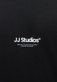 Fekete szövet, fehér hímzett szöveggel: "JJ Studios" és alatta kisebb betűkkel. Simább textúra, laza, hétköznapi dizájn.