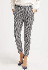 Vrouw in slim-fit broek met zwart-wit geometrisch patroon, witte blouse met lange mouwen en marineblauwe pumps met puntige neuzen, staat met de handen in de zakken.