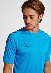 Koszulka sportowa z krótkim rękawem w jasnoniebieskim kolorze, z czarnymi akcentami na ramionach i logo na klatce piersiowej. Gładka tekstura, dopasowany krój.