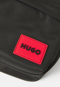 Schwarze strukturierte Stoffoberfläche mit einem roten rechteckigen Aufnäher, der mit erhabenen schwarzen Buchstaben "HUGO" beschriftet ist.