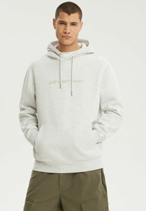 Młody mężczyzna w jasnoszarej bluzie z kapturem z logo "diverse" i oliwkowych spodniach typu cargo, ręce w kieszeniach bluzy, neutralne tło.