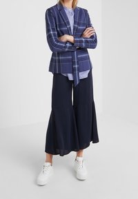 Blazer a quadri navy con laccetto, indossato sopra una camicia azzurro chiaro, abbinato a pantaloni navy a zampa d'elefante e sneakers bianche con zeppa.