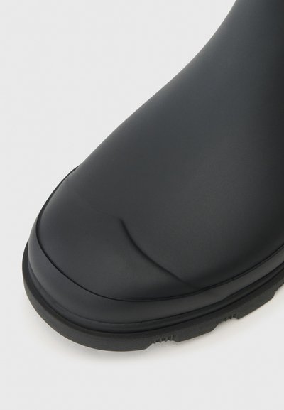 Zatvoreni kadar crne gumene čizme s teksturiranim potplatom na bijeloj pozadini.