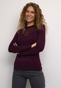 Maglietta viola a maniche lunghe con sovrapposizione trasparente all'escollatura, abbinata a jeans grigi con decorazioni di strass vicino all'orlo.