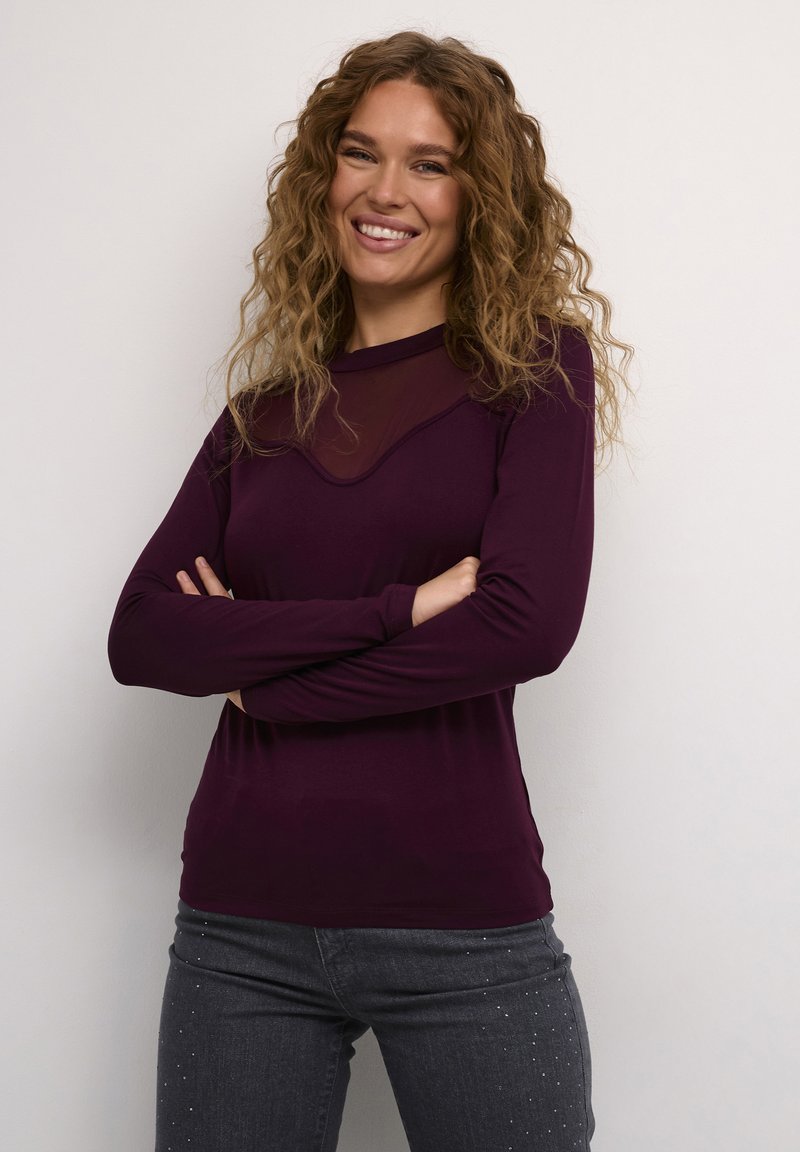 Maglietta viola a maniche lunghe con sovrapposizione trasparente all'escollatura, abbinata a jeans grigi con decorazioni di strass vicino all'orlo.