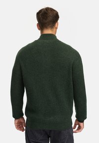 Dunkelgrüner, gerippter Pullover mit hohem Kragen, strukturiertem Stoff, langen Ärmeln und lockerer Passform. Sichtbare Nahtdetails am Saum.