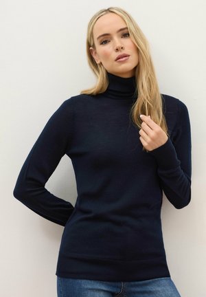 Femme aux longs cheveux blonds portant un pull à col roulé bleu marine et un jean bleu, debout contre un mur blanc uni.