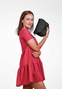 Černá látkov�á taška s hladkým povrchem, s zakřiveným designem a zipovým uzávěrem, držena osobou v červených šatech na bílém pozadí.