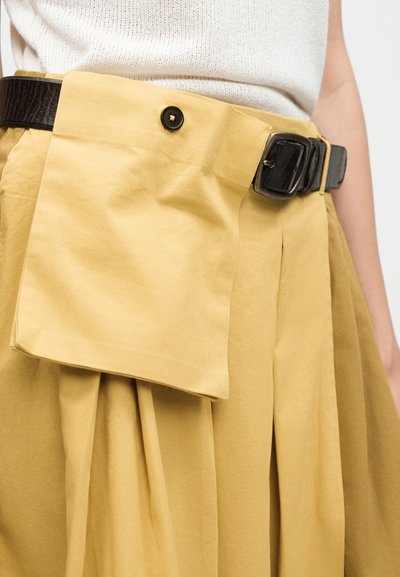 Γυναίκα φοράει μπεζ πλισέ φούστα με προσαρτημένη μεγάλη μπροστινή τσέπη και μαύρη δερμάτινη ζώνη, σε συνδυασμό με λευκό πλεκτό τοπ.