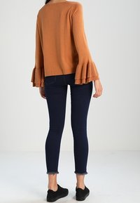 Roestkleurige trui met wijde mouwen, gecombineerd met donkerblauwe skinny jeans en zwarte instappers. Gladde textuur en comfortabele pasvorm.