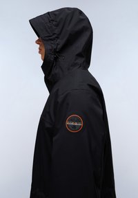 Svart vattentät jacka med huva, justerbar dragsko och ett cirkulärt logopatch med orange detaljer på ärmen.