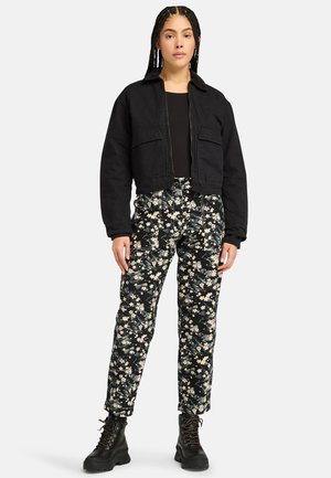 Black cropped jacket with two chest pockets, paired with floral-print black and white pants. Model wears black lace-up boots.