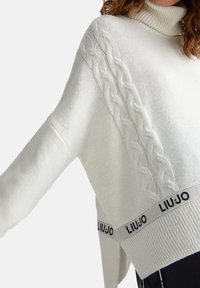 Witte gebreide trui met hoge kraag, voorzien van kabelbreisel details op de schouders en een gebrandmerkte tailleband met "LIU JO" in het zwart.