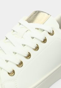 Λευκό αθλητικό παπούτσι με λείο, συνθετικό δερμάτινο επάνω μέρος, χρυσά μεταλλικά οπές για κορδόνια και μεταλλική χρυσή λεπτομέρεια στη φτέρνα. Σχέδιο με κορδόνια.