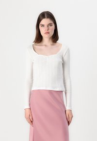 Chemise blanche à manches longues avec un décolleté carré et un motif texturé en côtes. Associée à une jupe en satin rose uni.