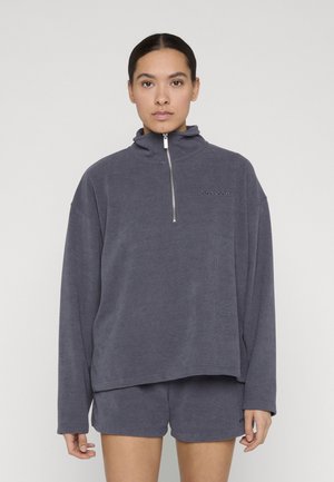 Donna con capelli scuri, indossa un pullover grigio in pile a maniche lunghe con colletto alto e pantaloncini grigi abbinati, in piedi davanti a uno sfondo semplice.