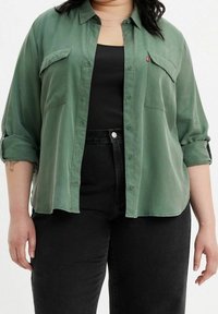 Chemise vert boutonnière avec une texture lisse, deux poches poitrine et des manches retroussées. Portée sur un débardeur noir et un jean noir taille haute.