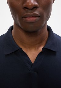 Tengerészkék pólóing, texturált pamut anyagból, V-nyakkivágással és klasszikus gallérral, sima varrással a nyakkivágás körül.