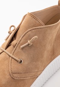 Chaussure en suede beige avec un bout arrondi, un système de lacets plats et des œillets métalliques. Semelle en caoutchouc blanche avec des détails texturés. Doublure intérieure lisse.