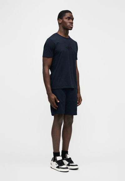 Tmavě modré bavlněné tričko s krátkými rukávy a logem na hrudi, doplněné tmavě modrými šortkami a černo-bílými teniskami.