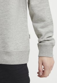 Grey sweatshirt with ribbed cuffs and hem, made of soft fabric. Features a small brand tag on the side. Visible hand and black pants.