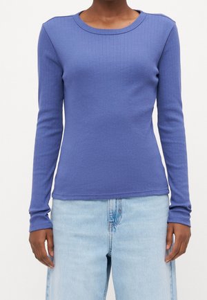 Osoba ubrana w dopasowaną, długorękawową fioletową prążkowaną koszulkę w połączeniu z jasnoniebieskimi dżinsami, ręce swobodnie opadające wzdłuż ciała.