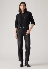 Zwarte spijkerblouse met drukknoopjes, twee borstzakken en opgerolde mouwen, gecombineerd met hoge zwarte spijkerbroek en zwarte enkellaarsjes.