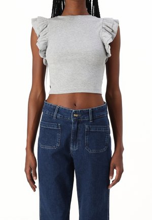 Kobieta ubrana w szary, bezrękawnikowy krótki top z falbankami na ramionach oraz ciemnoniebieskie, wysokie spodnie dżinsowe z przednimi kieszeniami na naszywki.