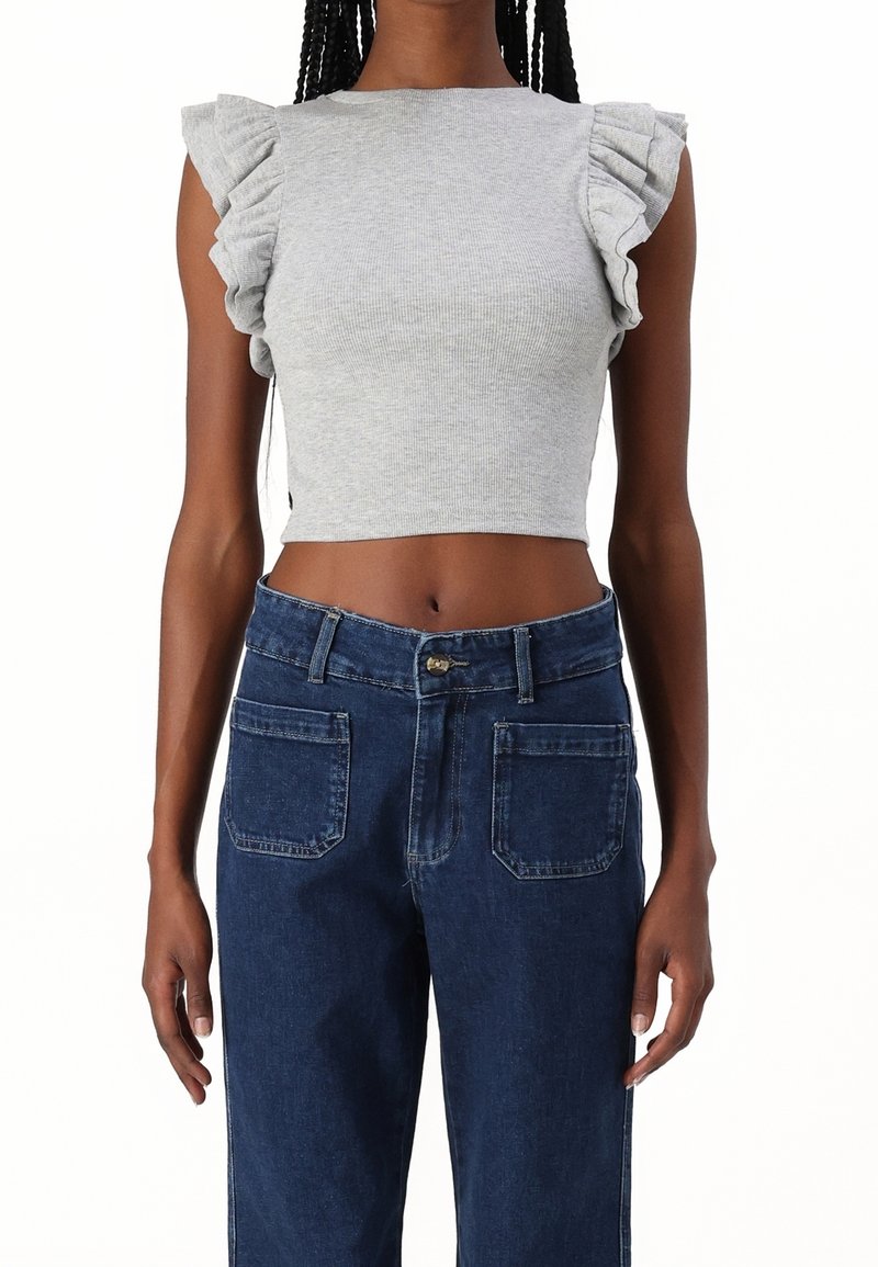 Femme portant un haut court gris sans manches avec des épaules à volants et un jean taille haute bleu foncé avec des poches plaquées à l'avant.