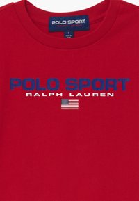 Červené bavlněné tričko se modrým logem "POLO SPORT", bílým textem "RALPH LAUREN" a malou grafikou americké vlajky níže.