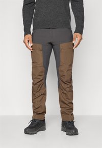 Man in een donkergrijze en bruine cargo broek met knoopzakken op de bovenbenen en zwarte wandelschoenen tegen een effen achtergrond.
